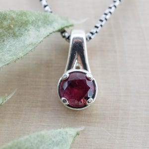 May include: A silver pendant necklace featuring a round, faceted deep red gemstone set in a four-prong silver mounting. The pendant hangs from a silver box chain, showcasing a polished metallic finish and a vibrant, dark red stone centre.