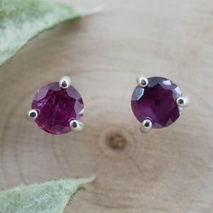 Op de afbeelding: Een paar oorknopjes met ronde, gefacetteerde diep magenta edelstenen. Elke steen wordt vastgehouden door drie zilverkleurige metalen pootjes in een driehoekige zetting, met een briljant slijpvorm die het licht op de donkerroze edelstenen weerkaatst.