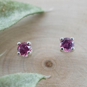May include: A pair of stud earrings featuring round, faceted, deep pink gemstones set in four-prong silver metal settings. The stones are circular and display a vibrant magenta hue, securely held by the metallic prongs in a classic stud design.