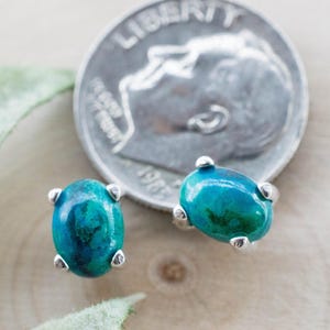 May include: A pair of oval-shaped turquoise and silver stud earrings. The earrings have a vibrant blue-green color and are set in silver. A U.S. dime is next to the earrings for size comparison. The earrings are on a wooden surface.