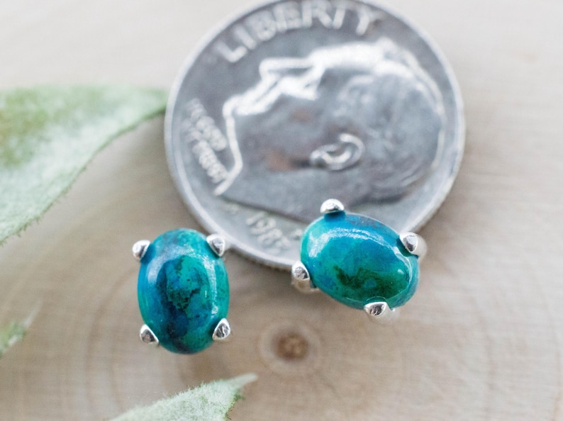 May include: A pair of oval-shaped turquoise and silver stud earrings. The earrings have a vibrant blue-green color and are set in silver. A U.S. dime is next to the earrings for size comparison. The earrings are on a wooden surface.
