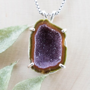 May include: A silver pendant necklace featuring a geode slice. The geode has a dark purple crystal center, surrounded by brown and green bands. The pendant is set in a silver bezel and hangs from a silver chain.
