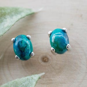 May include: A pair of oval-shaped stud earrings. Each earring features a vibrant teal and blue gemstone set in a silver metal prong setting. The earrings are displayed on a light wood surface.