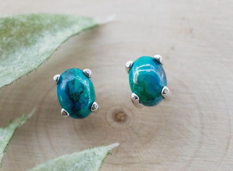 May include: A pair of oval-shaped stud earrings. Each earring features a vibrant teal and blue gemstone set in a silver metal prong setting. The earrings are displayed on a light wood surface.