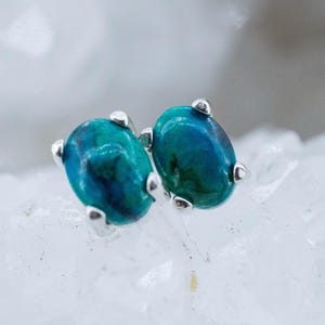 May include: A pair of oval-shaped stud earrings. Each earring features a vibrant, teal-colored stone with darker blue and green hues. The stones are set in a silver-colored metal with three small prongs securing each stone. The earrings are displayed on a white, textured surface.
