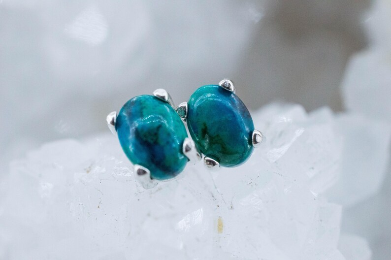 May include: A pair of oval-shaped stud earrings. Each earring features a vibrant, teal-colored stone with darker blue and green hues. The stones are set in a silver-colored metal with three small prongs securing each stone. The earrings are displayed on a white, textured surface.