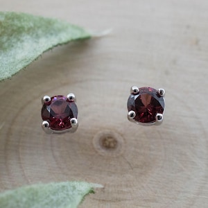 May include: A pair of stud earrings featuring round, deep red gemstones set in silver-colored metal. The earrings are displayed on a light wooden surface, with a blurred green leaf in the background. The gemstones have a rich, dark color.