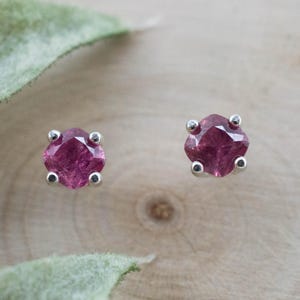 May include: A pair of stud earrings featuring faceted, cushion-cut, deep pink gemstones. Each stone is secured in a four-prong silver-toned metal setting. The earrings are displayed as individual pieces, showcasing the vibrant color and clarity of the stones.