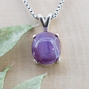 May include: A silver pendant necklace with a polished, oval purple gemstone. The gemstone is secured by four silver prongs. The necklace chain is a silver box chain. The background is a light brown surface with green leaves.