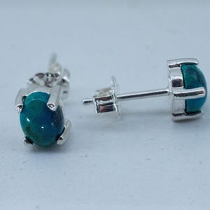 May include: A pair of stud earrings with a silver setting. Each earring features a round, polished, teal-colored stone. The earrings are displayed on a light gray surface, showcasing their design and color.
