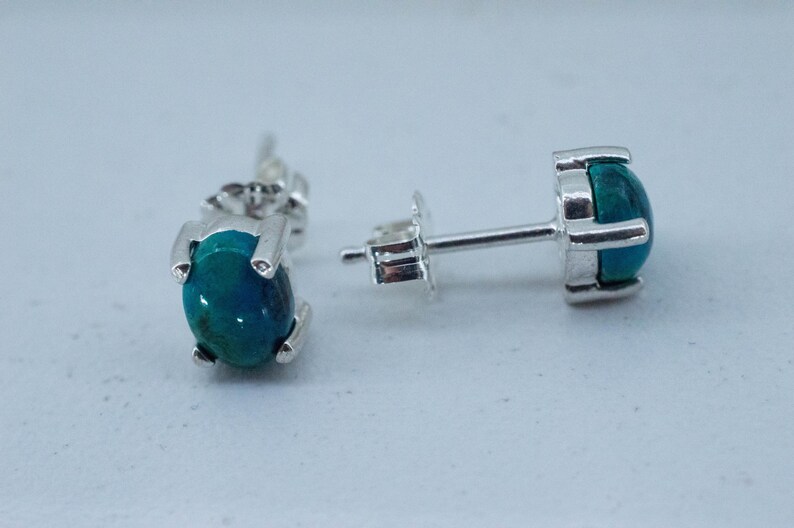 May include: A pair of stud earrings with a silver setting. Each earring features a round, polished, teal-colored stone. The earrings are displayed on a light gray surface, showcasing their design and color.
