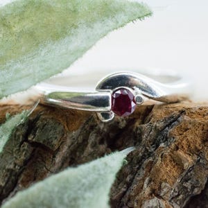 May include: A silver ring with a deep red gemstone, set on a textured wooden surface. The ring has a curved band and a central stone. A green leaf is in the foreground. The ring is likely made of silver.