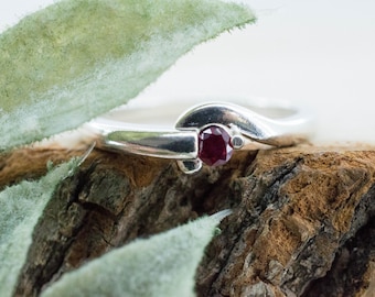 May include: A silver ring with a dark red gemstone, set on a textured wooden surface. The ring's design features a curved band and a prominent central stone. A green leaf is in the foreground.
