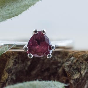May include: A silver ring featuring a pear-shaped, deep red gemstone held in place by a five-prong setting. The band of the ring is smooth and metallic, showcasing a polished finish that reflects light across its surface.