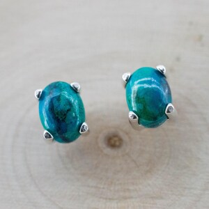 May include: A pair of oval-shaped stud earrings. Each earring features a vibrant, teal-colored stone with darker blue-green marbling. The stones are set in a silver-colored metal with four prongs. The earrings are displayed on a light wood surface.