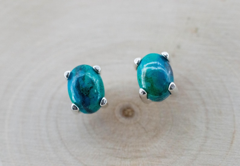 May include: A pair of oval-shaped stud earrings. Each earring features a vibrant, teal-colored stone with darker blue-green marbling. The stones are set in a silver-colored metal with four prongs. The earrings are displayed on a light wood surface.