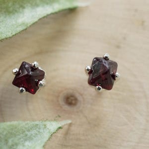 May include: A pair of silver stud earrings with small, square, red gemstone accents.