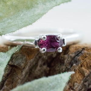 May include: A silver ring with a deep magenta oval gemstone. The ring is set on a textured wooden surface, with green leaves in the foreground. The gemstone is held in place by four silver prongs.