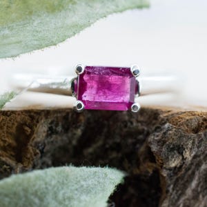 May include: A silver ring featuring a rectangular, emerald-cut vibrant pink gemstone set in a four-prong setting. The band is smooth and polished, showcasing a clean, metallic finish that contrasts with the deep, saturated color of the stone.