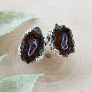 May include: A pair of stud earrings featuring a natural geode design. The earrings have a dark green outer edge with a brown and white center. The earrings are set in silver metal with small, round accents around the edges.