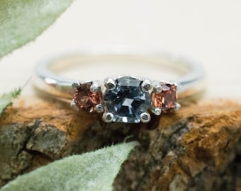 May include: A silver ring with three gemstones. The center stone is a large, dark blue, faceted gemstone. The two smaller stones on either side are a reddish-brown color.