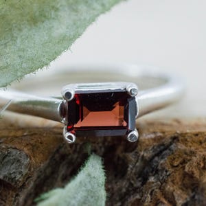 May include: A silver ring featuring a rectangular, dark red gemstone. The ring's band is smooth and polished, with the gemstone held in place by four small prongs. The ring is set against a natural wood background.