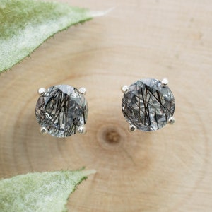 May include: A pair of round stud earrings. Each earring features a clear, faceted gemstone with black inclusions, set in a silver-colored metal. The earrings are displayed on a light-colored wooden surface, with green leaves in the background.