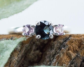 May include: A silver ring with three gemstones. The center stone is a dark blue, flanked by two smaller, light pink stones. The ring is set against a natural wood and green leaf background.
