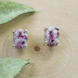 Puede incluir: Un par de pendientes de botón de forma ovalada. Cada pendiente presenta una piedra moteada rosa y blanca engastada en un bisel de metal plateado. Los pendientes se exhiben sobre una superficie de madera con hojas verdes.