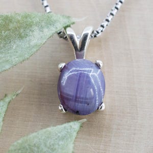 May include: A silver pendant necklace with a large, oval purple gemstone. The gemstone is secured by four silver prongs and hangs from a silver chain. The background includes green leaves and a light-coloured wooden surface.