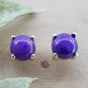 May include: A pair of round, purple stud earrings with silver prongs. The earrings are set against a light wooden surface, with a blurred green leaf in the background. The earrings are a simple, elegant design.