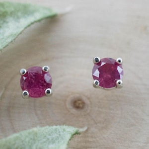 May include: A pair of round, faceted, deep pink gemstone stud earrings set in four-prong silver metal settings. Each earring features a circular stone with a brilliant cut, securely held by four small silver prongs that frame the vibrant pink gem.