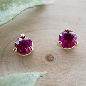 May include: A pair of stud earrings featuring round, faceted, deep pink gemstones set in gold-colored metal. The earrings are displayed on a light-colored wooden surface, with a green leaf in the background.