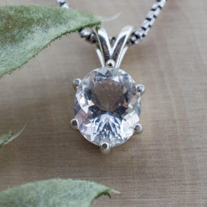 May include: A clear, oval-cut gemstone pendant set in a polished silver prong setting. The pendant hangs from a silver box chain necklace. The faceted stone reflects light, showcasing a transparent, colourless appearance with a brilliant sparkle.