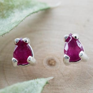 May include: A pair of stud earrings featuring vibrant, faceted ruby gemstones set in a silver metal. The ruby stones have a deep red hue and are mounted on a light-colored wooden surface.