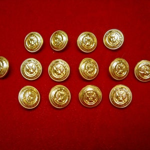 May include: A collection of gold-colored buttons with a detailed emblem design. The buttons are round with a raised edge and a decorative emblem in the center. The buttons are arranged on a red surface.