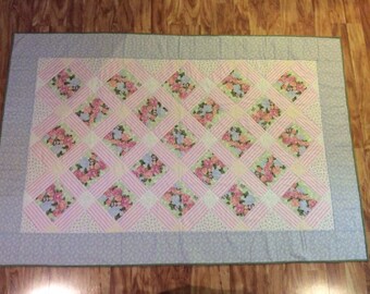 May include: A rectangular quilt with a floral pattern. The quilt features a light blue border, a central section with pink and white striped squares, and floral diamond patterns in pink, blue, and green.