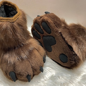 May include: A pair of brown furry bear paw slippers with black claws and paw pads.