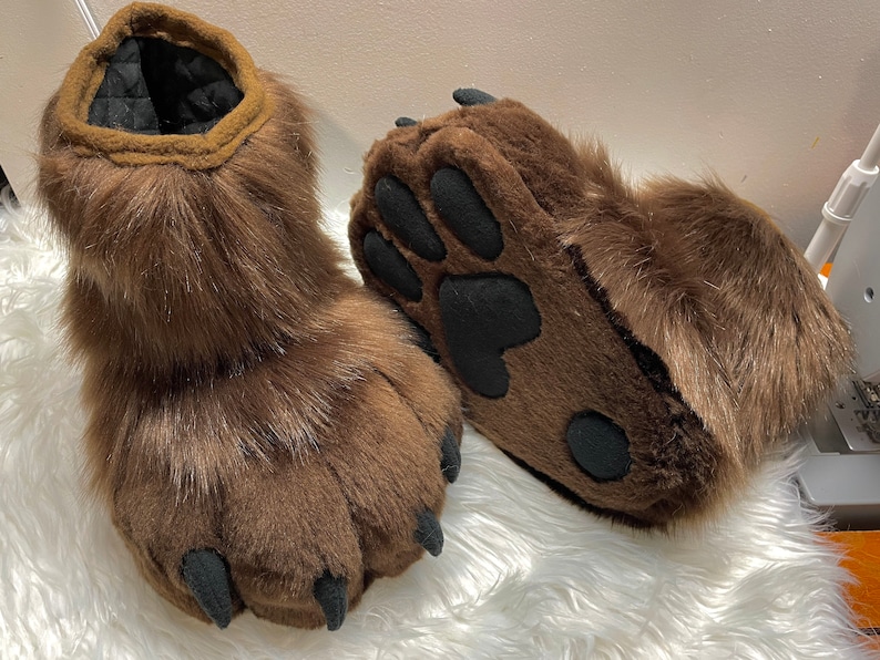 May include: A pair of brown furry bear paw slippers with black claws and paw pads.