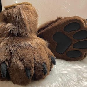 May include: A pair of brown furry bear paws with black claws and black paw pads. The paws are on a white fluffy surface.