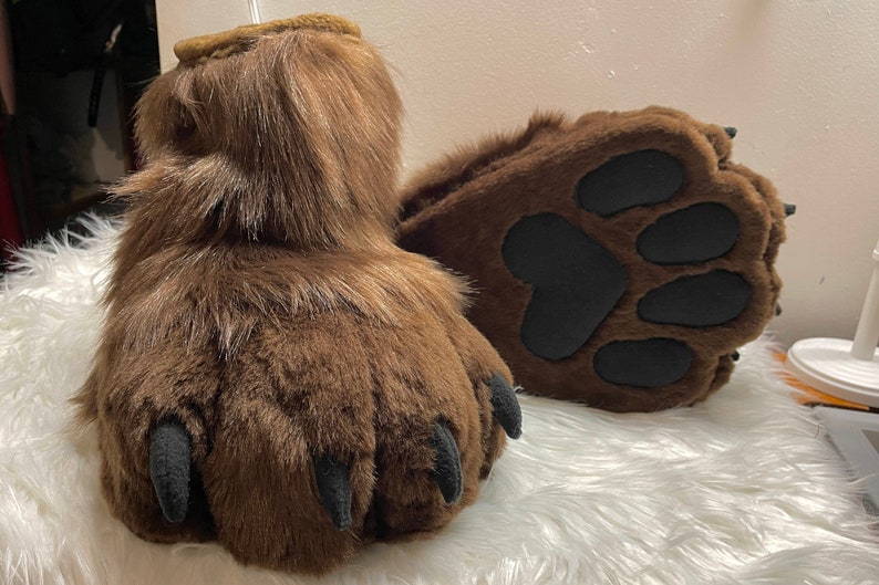 May include: A pair of brown furry bear paws with black claws and black paw pads. The paws are on a white fluffy surface.