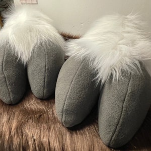 Puede incluir: Dos almohadas de felpa gris y blanca con forma de piernas. Las almohadas tienen una parte superior blanca peluda y una parte inferior gris.