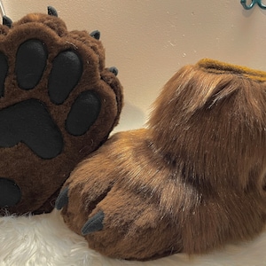 May include: A pair of brown furry paw-shaped slippers with black paw pads and claws. The slippers are made of soft, plush fabric and have a realistic paw design.