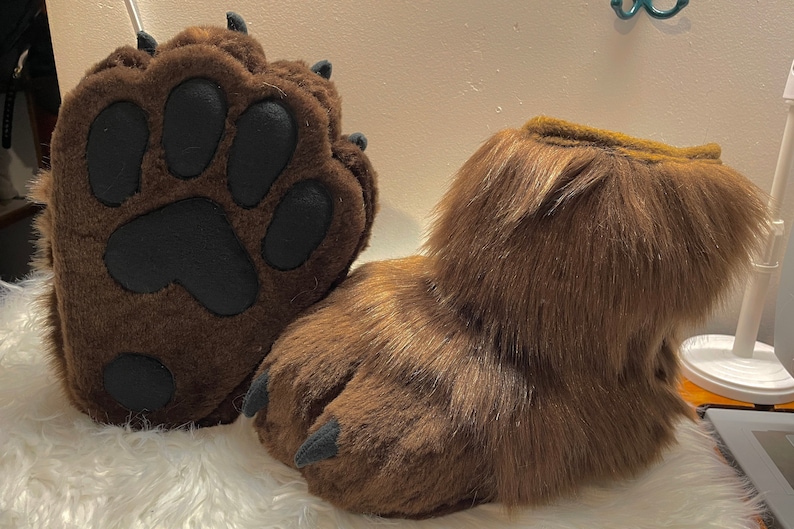 May include: A pair of brown furry paw-shaped slippers with black paw pads and claws. The slippers are made of soft, plush fabric and have a realistic paw design.