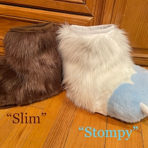 May include: Two pairs of fuzzy animal paw slippers. The brown pair is labeled "Slim" and the white pair with blue paw pads is labeled "Stompy".