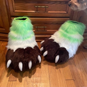 May include: A pair of furry brown and white paw-shaped slippers with green and white fur accents. The slippers have claws and are made of soft, plush fabric.
