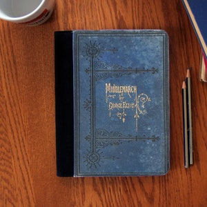 Può includere: Un quaderno blu con una rilegatura nera e lettere dorate. La copertina riporta la scritta "Middlemarch di George Eliot".