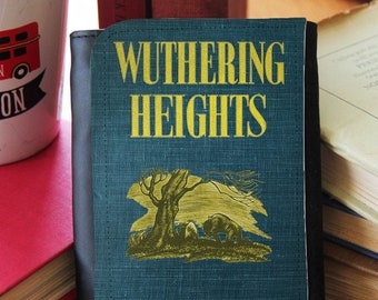 Könnte beinhalten: Eine blaue Stoffbörse mit schwarzem Rand. Die Vorderseite der Börse zeigt den Titel "Wuthering Heights" in gelber Schrift und eine Schwarzweißillustration eines Baumes und einer Figur im Hintergrund. Der Autor, "Emily Brontë", ist unten in Kursivschrift gedruckt.