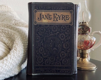 Könnte beinhalten: Ein schwarzes, ledergebundenes Buch mit goldenen Lettern, auf dem "Jane Eyre" und "Household Edition" steht. Das Cover zeigt ein Blumenmuster.