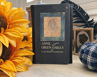 May include: A vintage hardcover book titled "Anne of Green Gables" by L.M. Montgomery. The book has a dark blue cover with a faded image of a young girl on the front. The book is sitting on a wooden surface with sunflowers and a plaid fabric box.